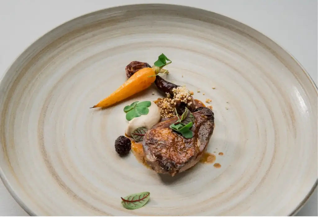Gourmet plated lamb with roasted carrot, puree, dried fruits, puffed grains, and microgreens garnish on ceramic plate