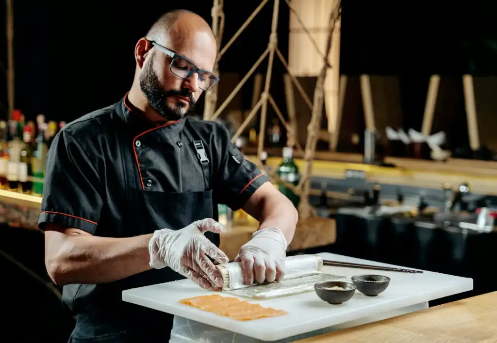 A focused chef in a black jacket, safety glasses, and latex gloves carefully rolling a preparation on a marble board, with sliced salmon and seasoning bowls alongside, in a dimly lit restaurant kitchen