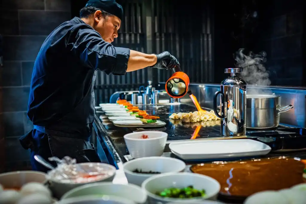 A chef in a dark uniform using a blowtorch to flame-finish dumplings on a professional kitchen counter, surrounded by multiple prepped ingredient bowls and a row of plated dishes, with steam rising in the background