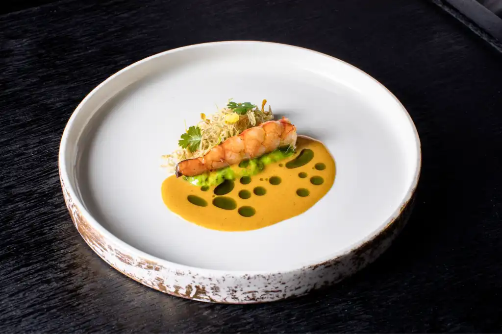 An elegantly plated fine dining dish featuring a seared prawn on green purée with golden bisque sauce dotted with herb oil, garnished with crispy frisée and microgreens, served on a rustic white ceramic plate against a dark surface