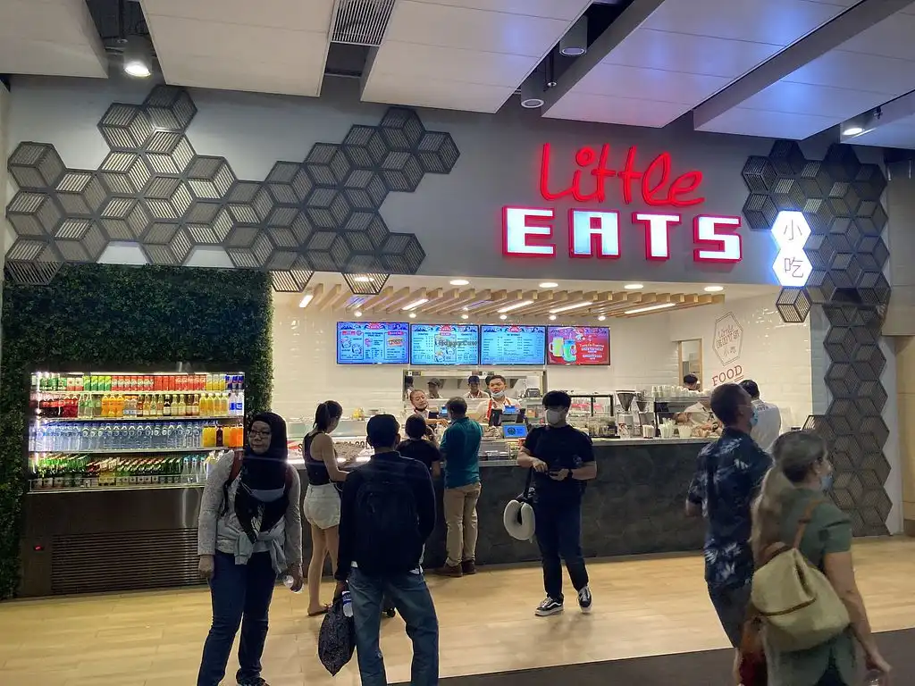 Busy Little Eats food stall at Ngurah Rai International Airport Bali, with a red neon sign, hexagonal wall décor, digital menu boards, a refrigerated drinks display, and travelers queuing at the counter