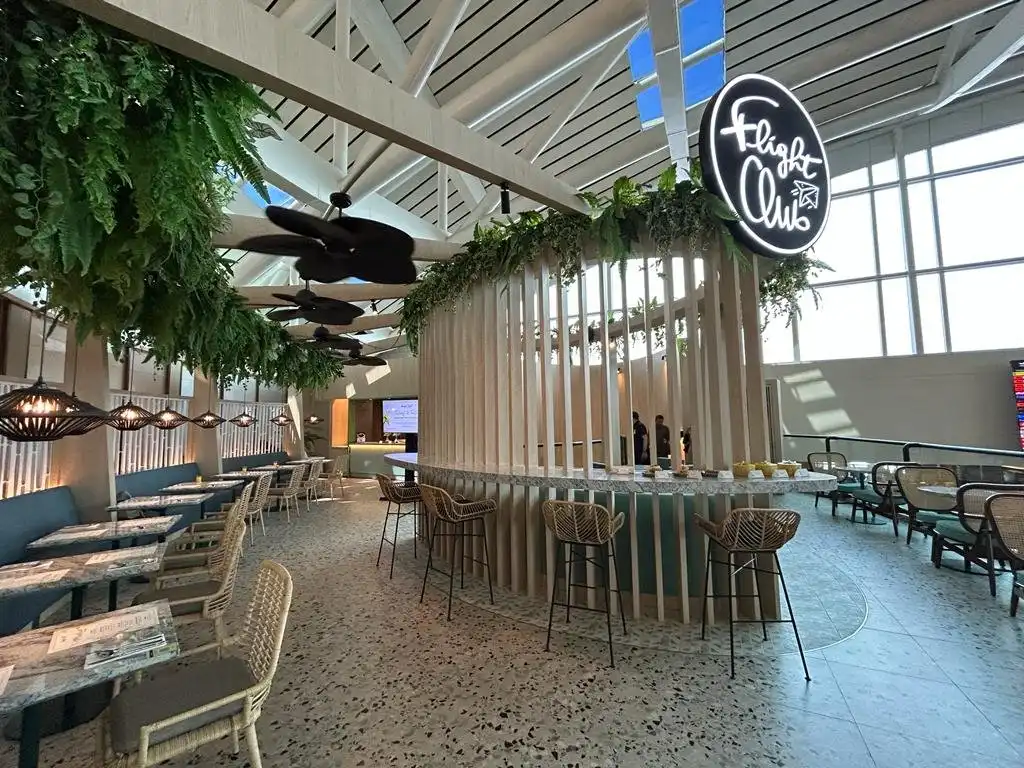 Interior of Flight Club restaurant at Ngurah Rai International Airport Bali, featuring a tropical-chic design with rattan bar stools, hanging greenery, ceiling fans, and a neon circular logo sign