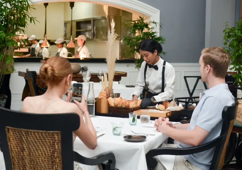 Fine dining table setting with server presenting bread selection to guests at elegant restaurant with open kitchen.