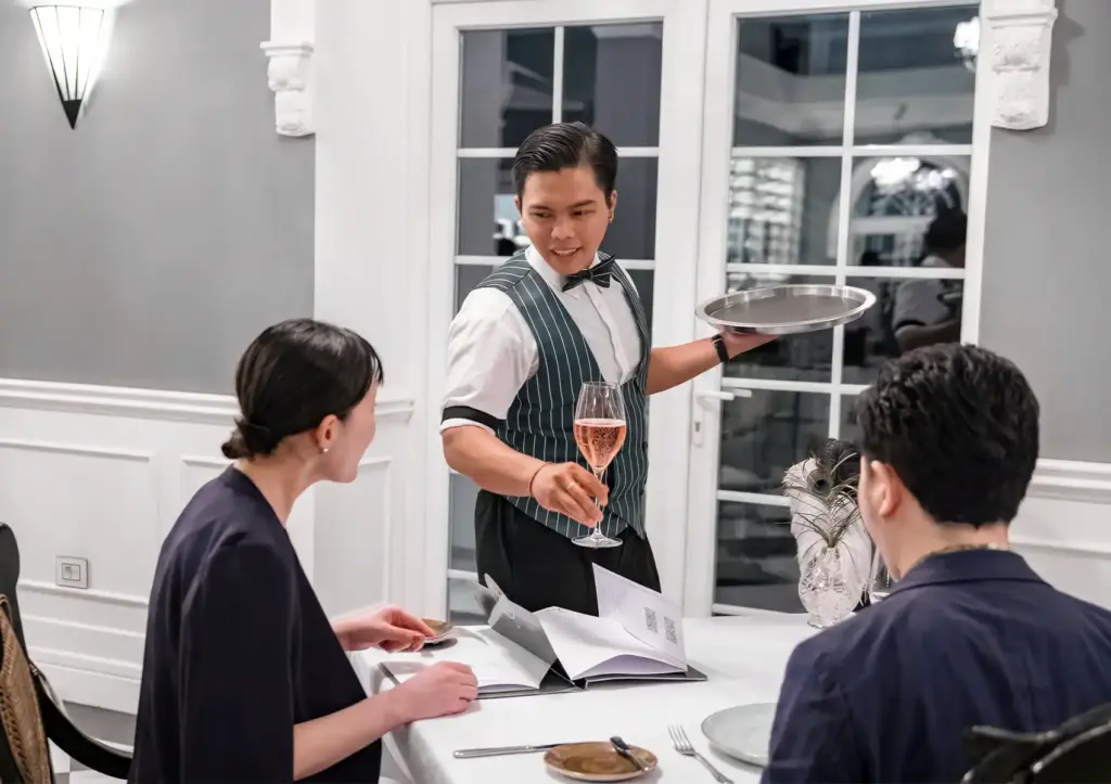 Server presenting rosé wine to diners at elegant restaurant table with menus and white table setting.