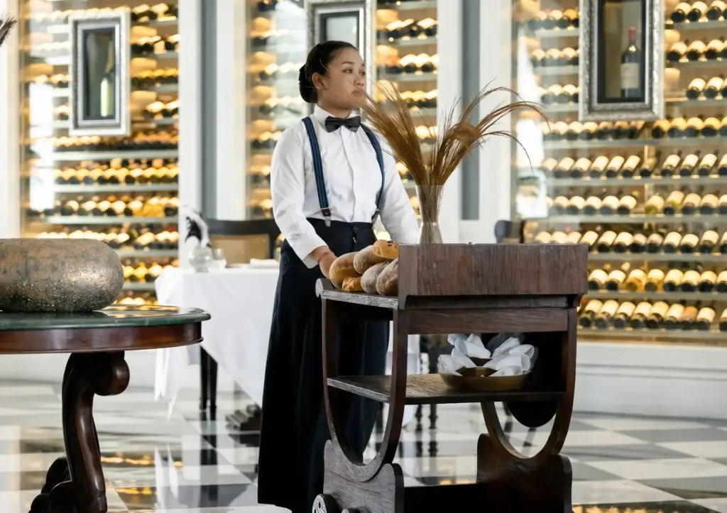 Elegant restaurant with server beside bread cart and wine display wall in refined dining atmosphere.