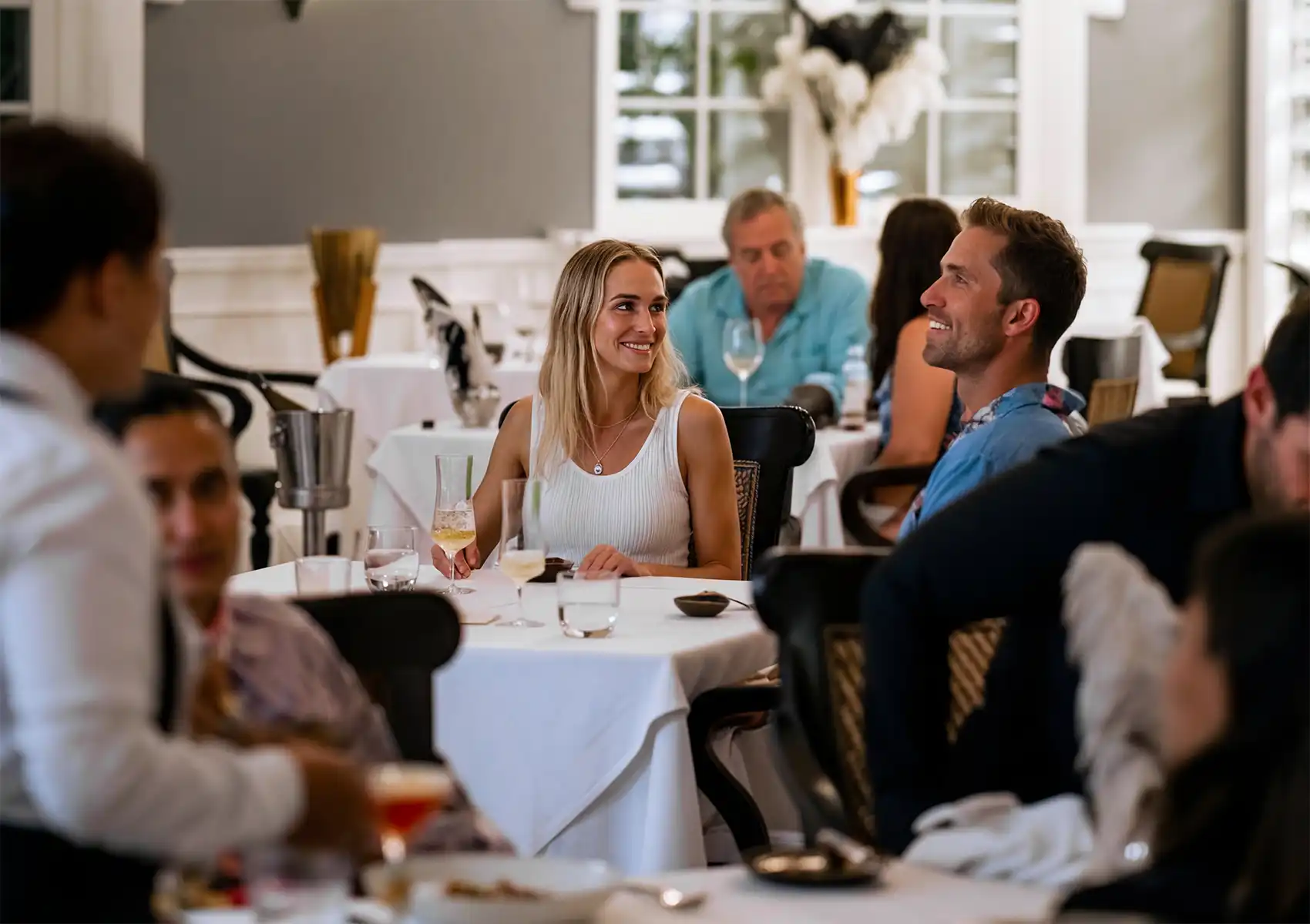 Elegant restaurant dining scene with guests enjoying wine and conversation at white‑tablecloth tables.