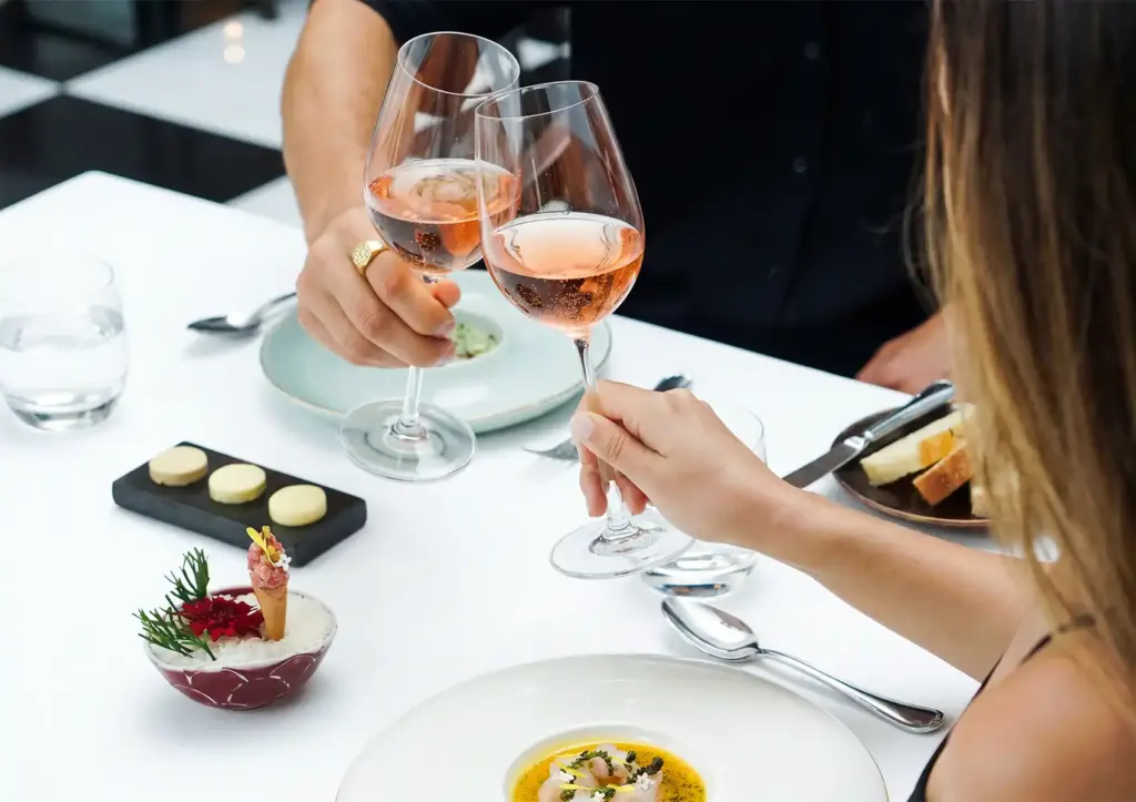 Fine dining celebration with guests clinking glasses of rosé wine over gourmet dishes on an elegant white‑tablecloth table.