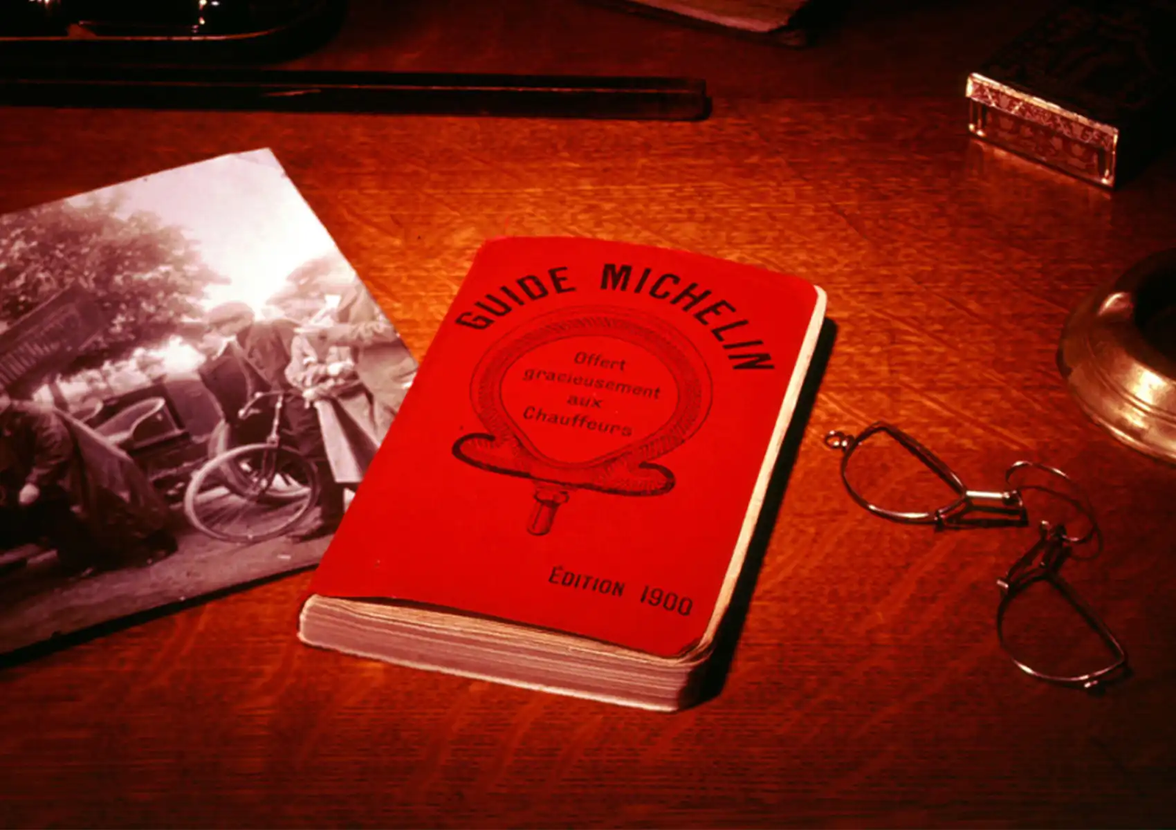 The iconic red-covered Guide Michelin Édition 1900 — inscribed "Offert gracieusement aux Chauffours" (offered free to motorists) — resting on a warm wooden desk beside a vintage black-and-white photograph of an early automobile and a pair of antique wire-rimmed spectacles
