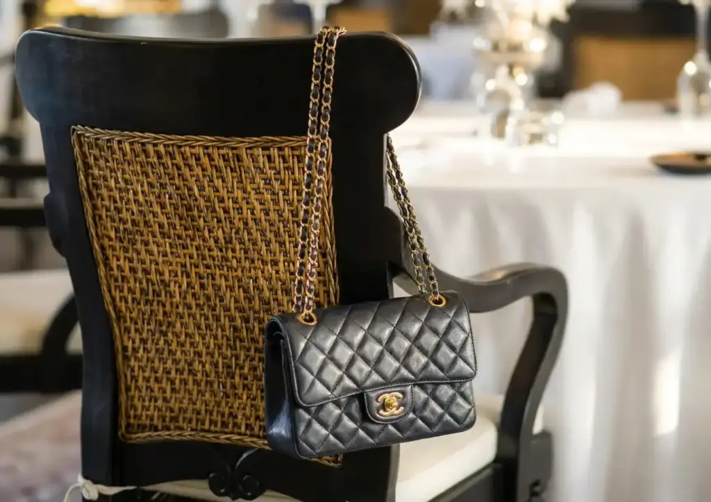 A quilted black leather handbag with gold chain straps and a gold double-C clasp resting on a woven rattan dining chair, with a white-clothed fine dining table and silverware softly blurred in the background
