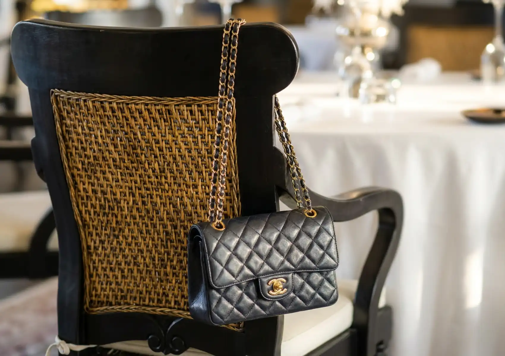 A quilted black leather handbag with gold chain straps and a gold double-C clasp resting on a woven rattan dining chair, with a white-clothed fine dining table and silverware softly blurred in the background