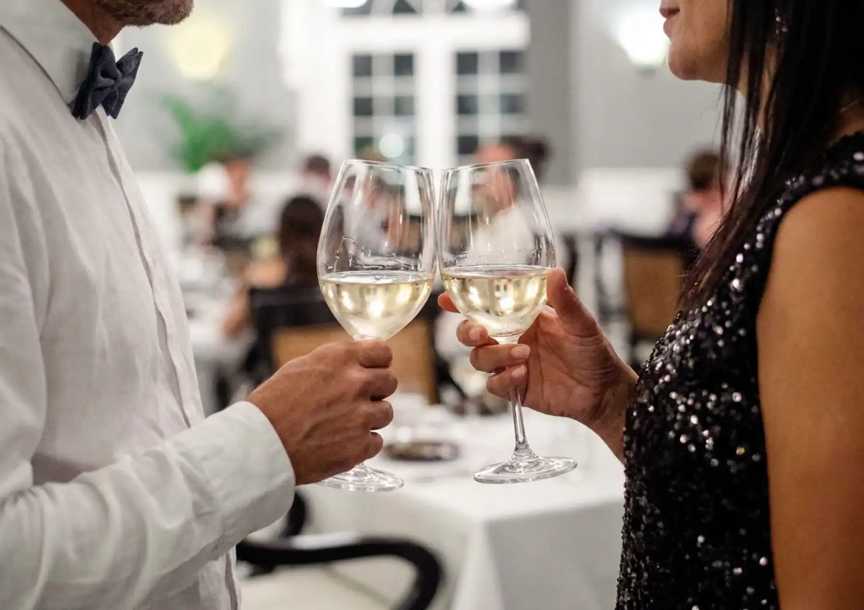A close-up of a couple clinking two glasses of white wine in a warmly lit fine dining restaurant, the man in a white shirt and bow tie and the woman in a black sequined dress, with white-clothed tables and other diners softly blurred in the background