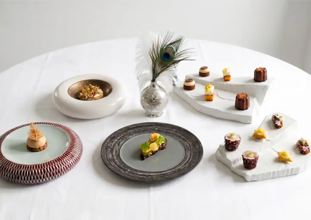 A fine dining tasting menu spread at Aperitif Restaurant Ubud, featuring multiple precisely plated courses displayed on artisan ceramics and tiered white marble stands, including petit fours, amuse-bouche bites, and elegantly composed main course plates, styled with a peacock feather centerpiece on a white linen table