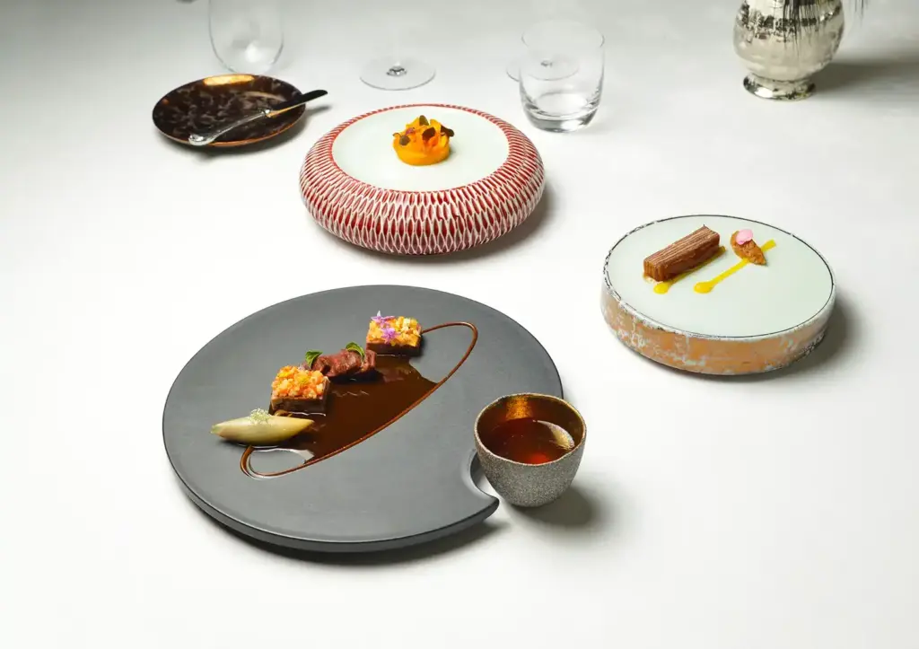 An overhead styled tablescape of three artfully plated fine dining courses on sculptural ceramic vessels — a crescent-shaped slate plate with seared beef medallions, edible flowers, and rich jus beside a small gold-rimmed cup; a red lattice-patterned drum plate with a delicate orange purée; and a celadon disc with a glazed terrine and saffron sauce — arranged on a white tablecloth with crystal glassware