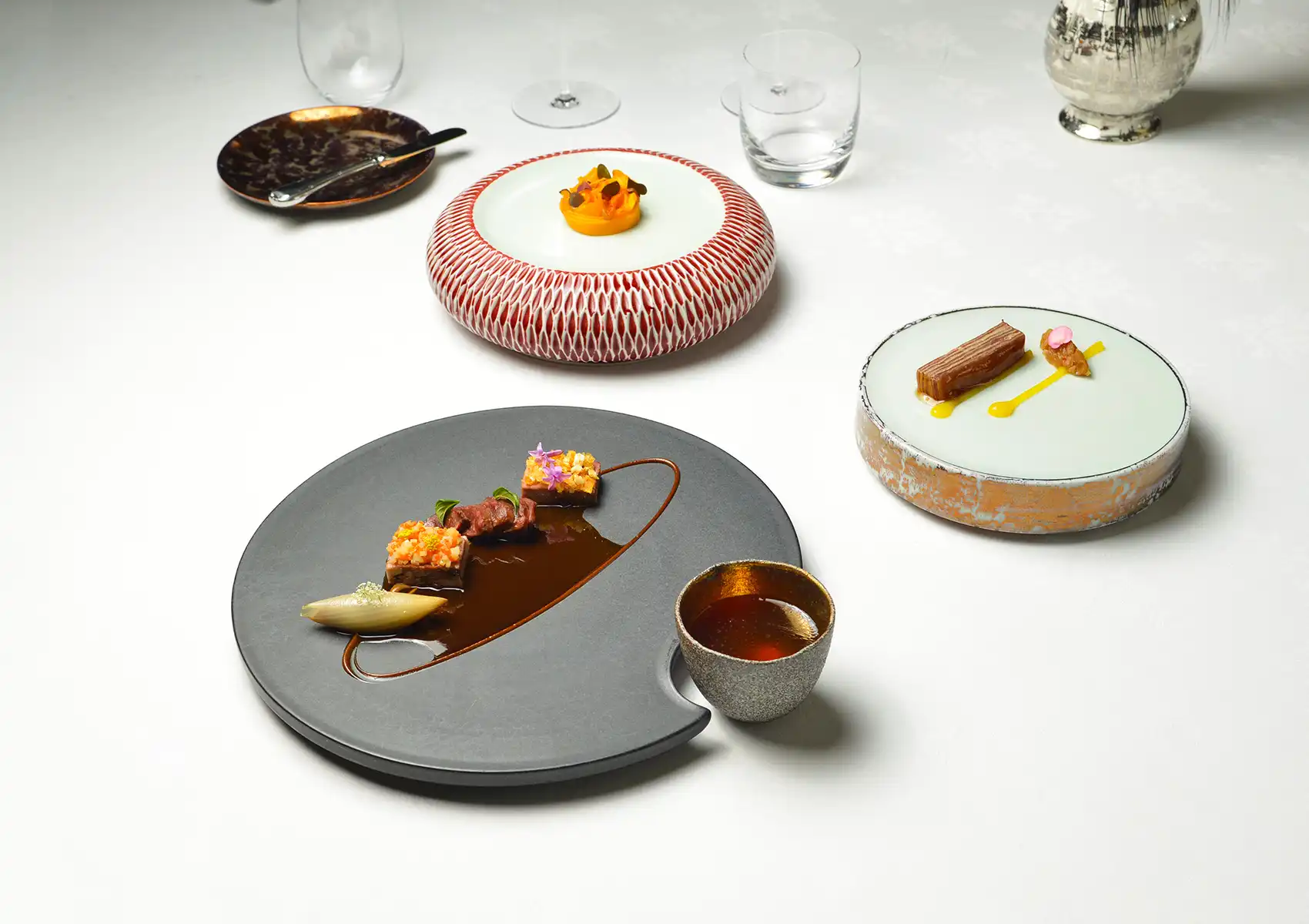 An overhead styled tablescape of three artfully plated fine dining courses on sculptural ceramic vessels — a crescent-shaped slate plate with seared beef medallions, edible flowers, and rich jus beside a small gold-rimmed cup; a red lattice-patterned drum plate with a delicate orange purée; and a celadon disc with a glazed terrine and saffron sauce — arranged on a white tablecloth with crystal glassware
