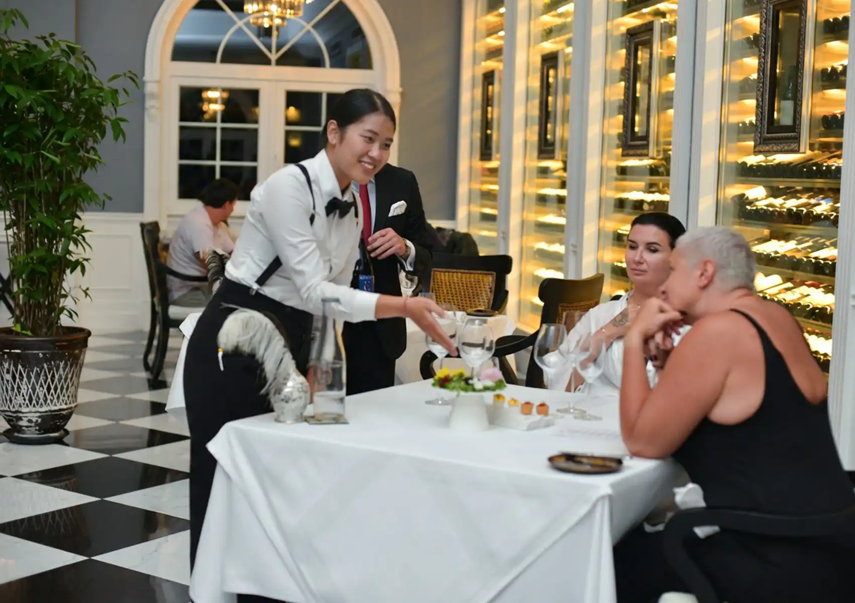 A smiling server in a white shirt, bow tie, and suspenders attentively presenting an amuse-bouche to two diners at a white-clothed table inside an elegant restaurant with black-and-white checkered floors, arched windows, and a dramatically backlit floor-to-ceiling wine cellar wall