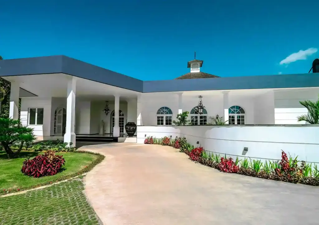The grand white colonial-style exterior of a fine dining restaurant, featuring a sweeping driveway lined with vibrant red tropical plants, a columned portico entrance with wrought iron lanterns, arched windows, and a cupola rooftop detail under a vivid blue sky