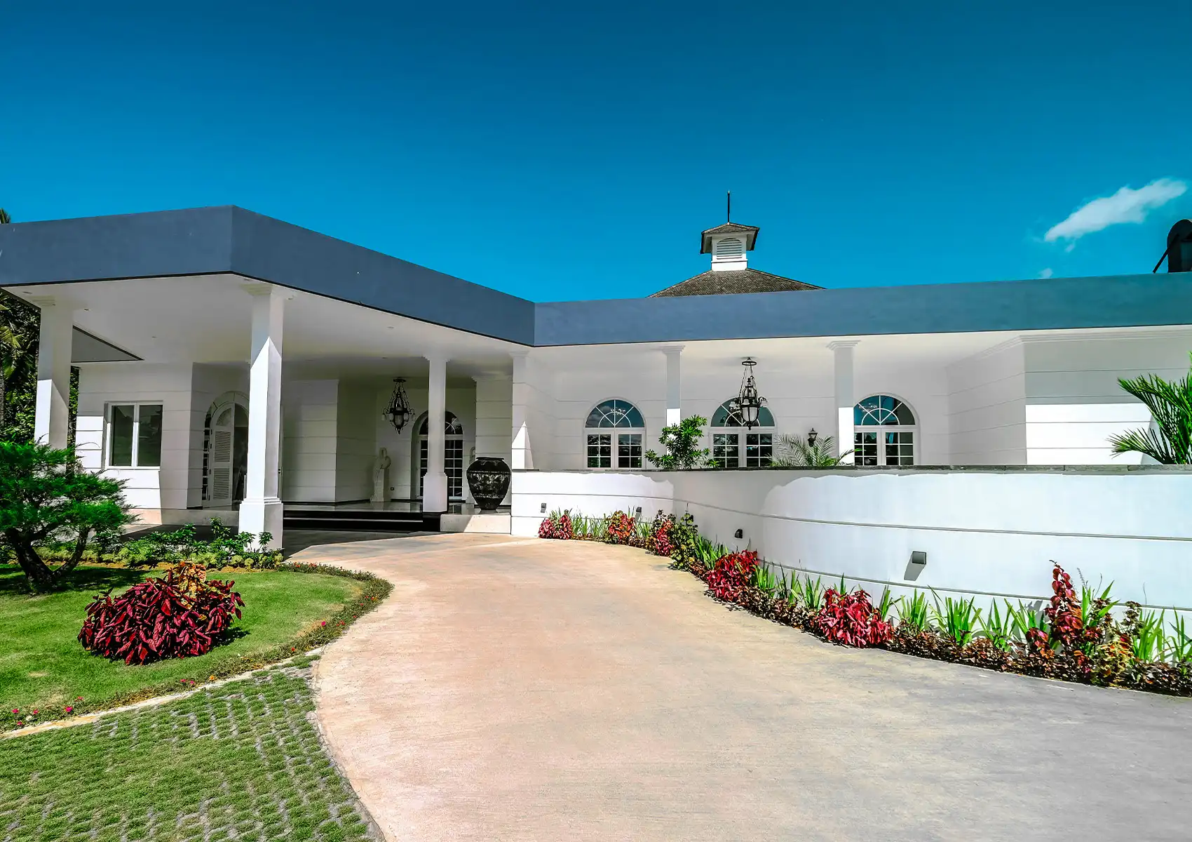 The grand white colonial-style exterior of a fine dining restaurant, featuring a sweeping driveway lined with vibrant red tropical plants, a columned portico entrance with wrought iron lanterns, arched windows, and a cupola rooftop detail under a vivid blue sky