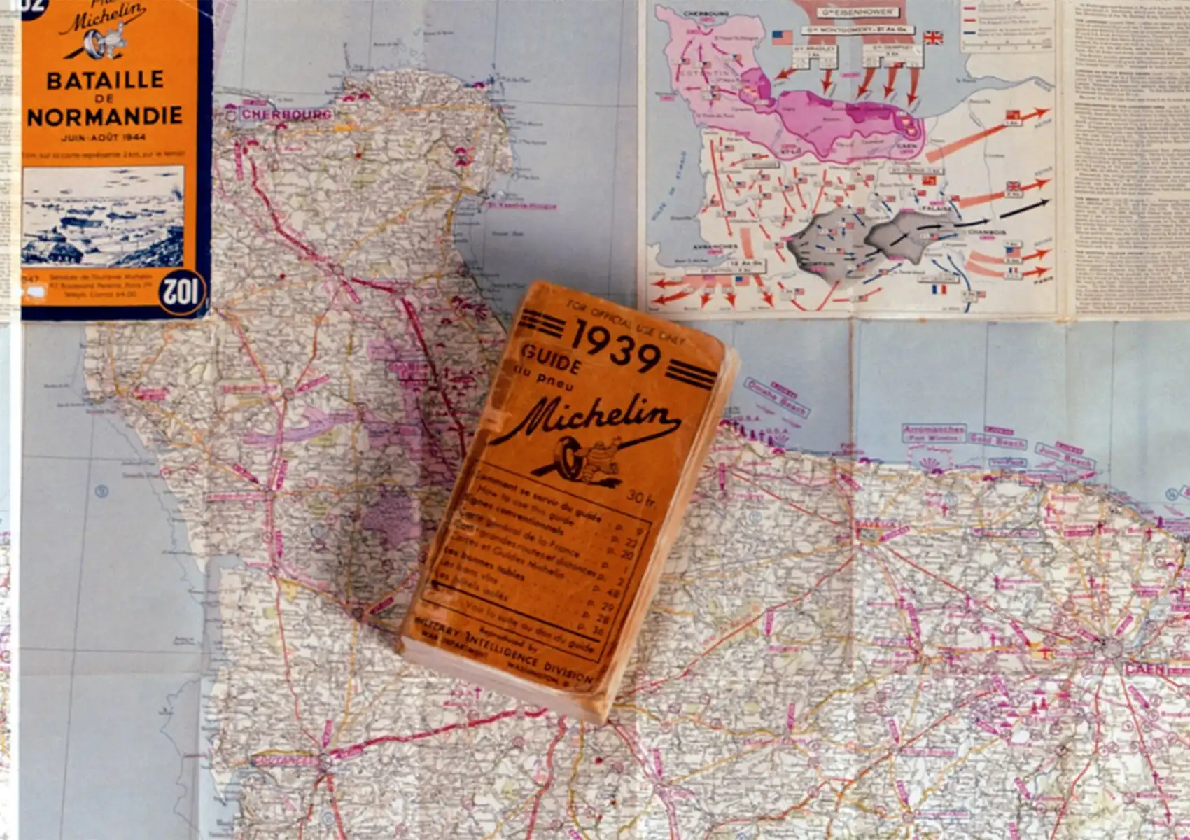 A 1939 Guide du Pneu Michelin booklet stamped "For Official Use Only" by the Military Intelligence Division, resting on a detailed WWII-era military map of Normandy, France, alongside an orange Michelin "Bataille de Normandie" map from June–August 1944