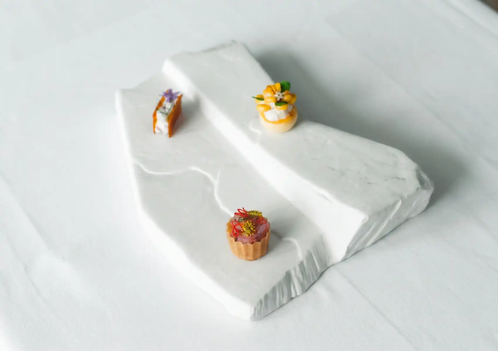 Three exquisite bite-sized amuse-bouche arranged on a sculptural tiered white stone platter — a glazed caramelised piece topped with purple flowers, a golden tartlet crowned with mango and white blossoms, and a fluted pastry cup filled with vibrant red ingredients and micro herbs