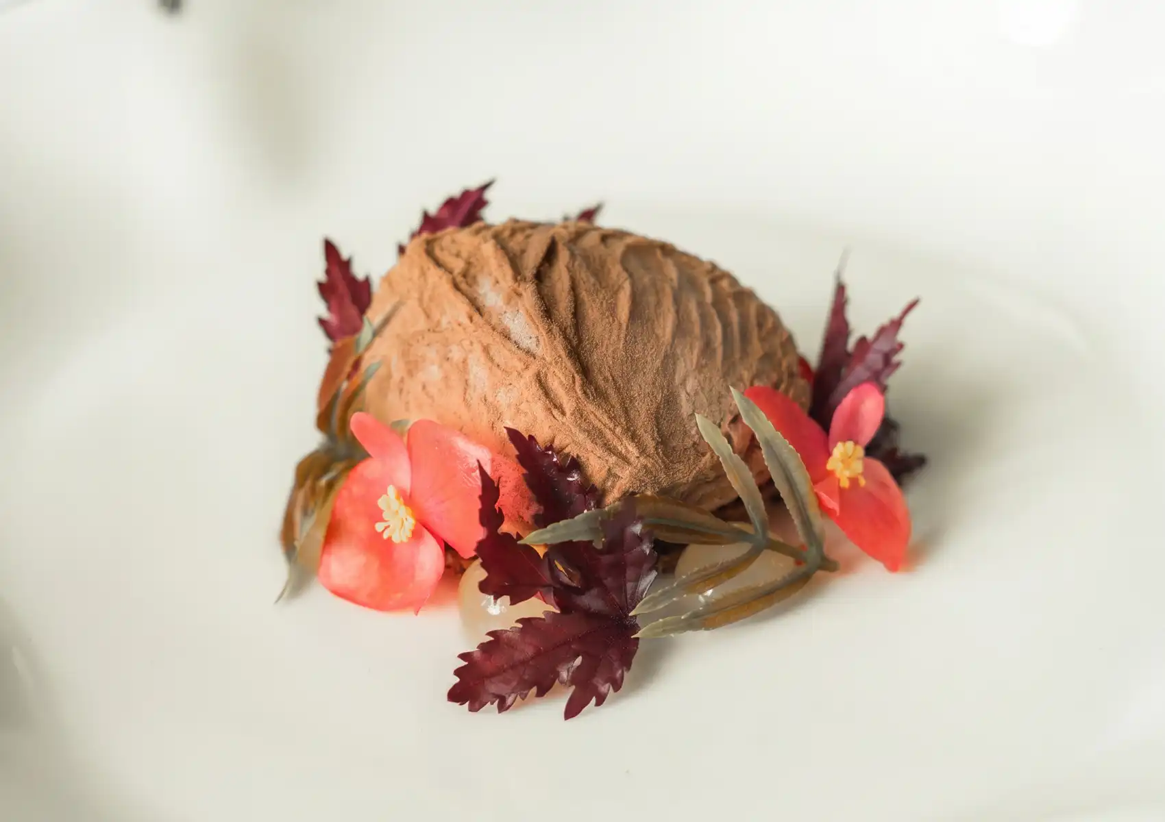 A stunning fine dining dessert presented on a white plate — a smooth domed chocolate mousse dusted with cocoa powder, encircled by vivid coral begonia blossoms, deep burgundy micro leaves, and slender herb sprigs, evoking an enchanted forest scene