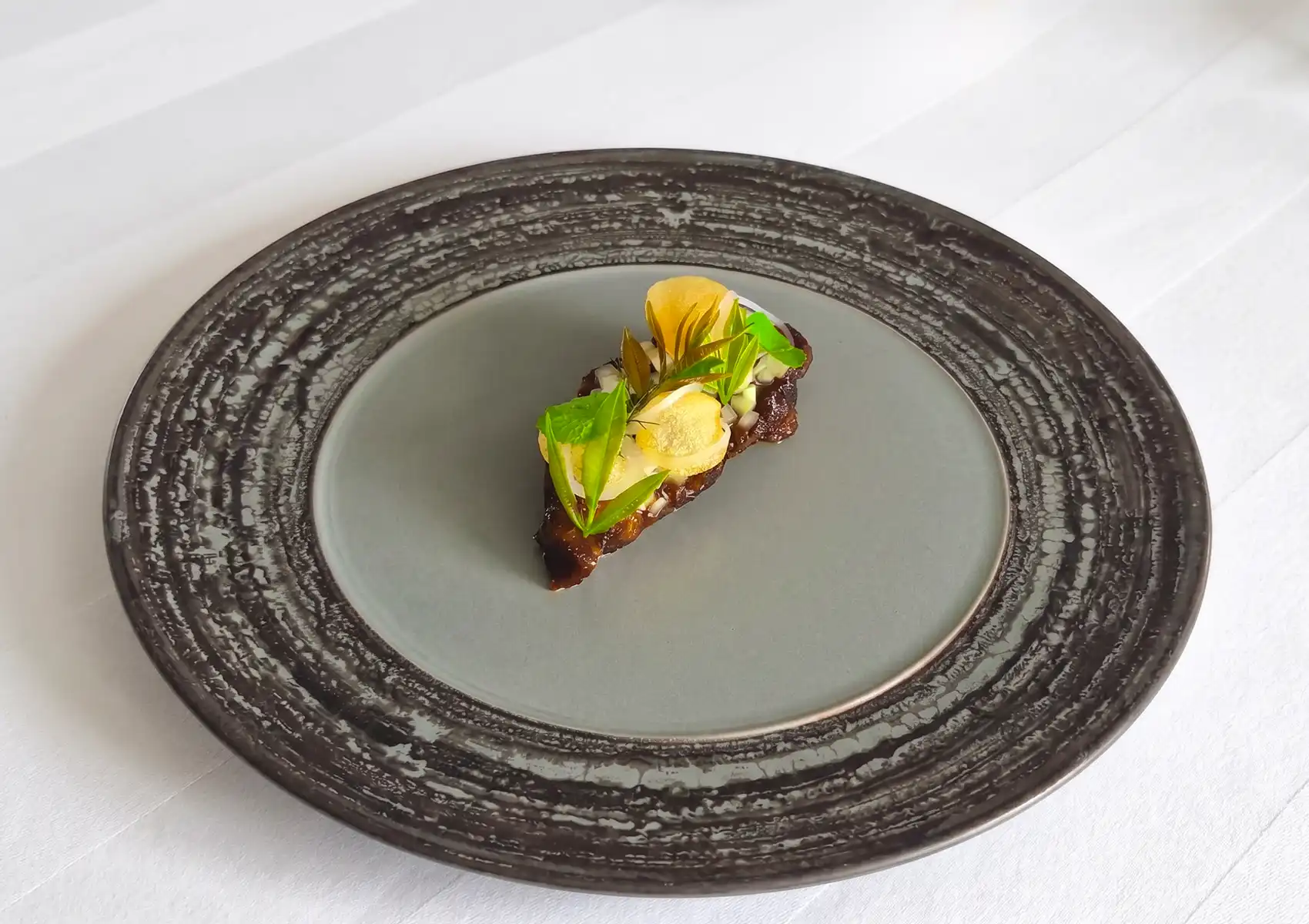 A beautifully plated dish showcasing Indonesian ingredients in fine dining, featuring a glazed piece of meat delicately topped with fresh green herbs and thin yellow crisps on a rustic, textured gray plate.