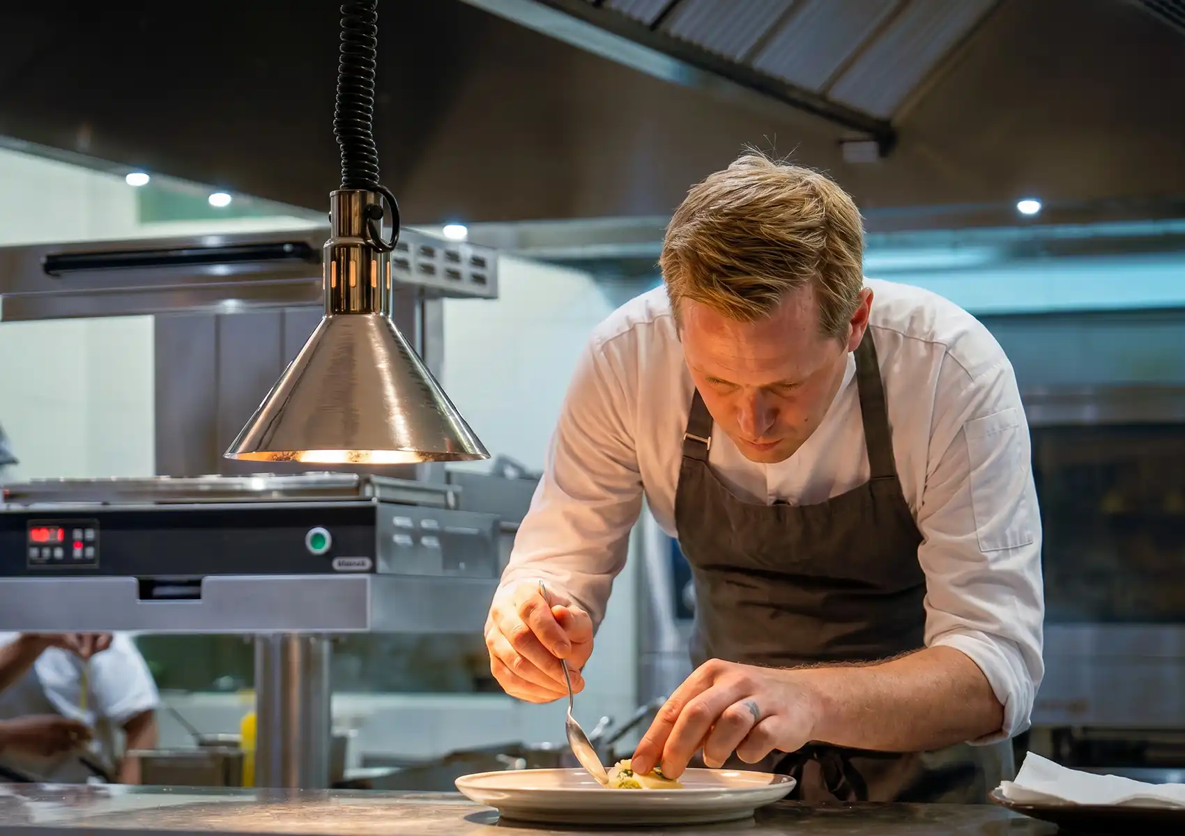 A professional chef in a grey apron meticulously plating a gourmet dish under a heat lamp in a modern kitchen, capturing the culinary dedication of fine dining Ubud.