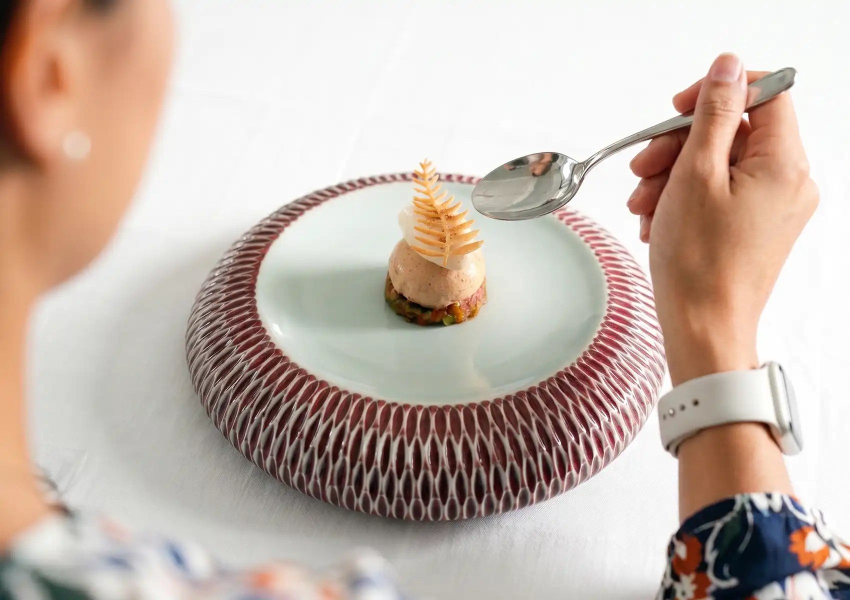 A guest holding a silver spoon about to enjoy an elegant, layered heirloom tomato appetizer on a textured artisan ceramic plate, experiencing sustainable fine dining Ubud.
