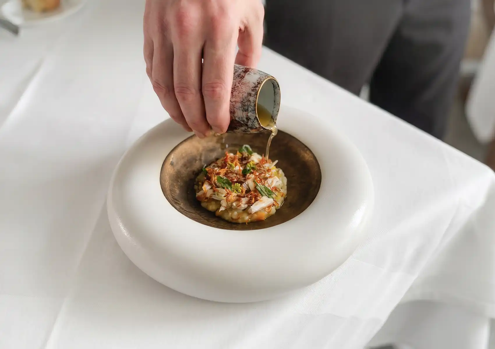 A close-up of a chef pouring a clear, savory broth from a small patterned ceramic pitcher over a meticulously arranged seafood and herb dish, highlighting the elegant use of Indonesian ingredients in fine dining.