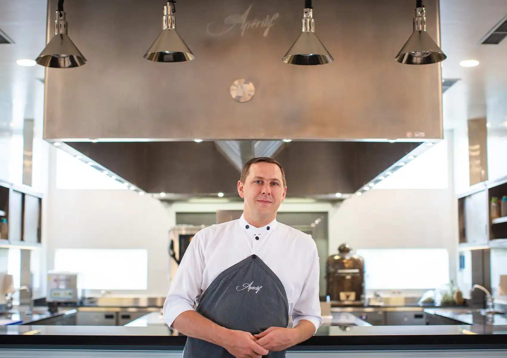 A professional chef wearing a white uniform and grey apron stands proudly in front of a state-of-the-art, stainless-steel open kitchen, ready to prepare a spectacular meal at a premier destination for Fine dining bali.