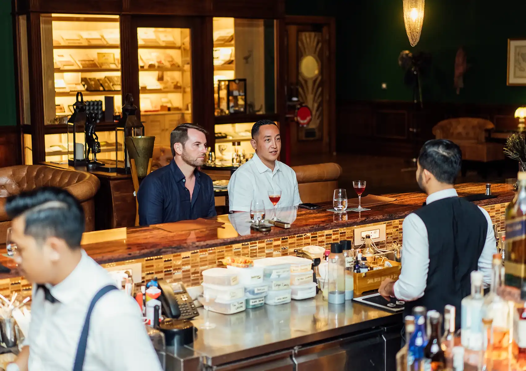 Two guests sit at an elegant, warmly lit speakeasy bar with a polished wood counter, chatting with the bartender while enjoying craft cocktails as a prelude to an exquisite Fine dining bali experience.