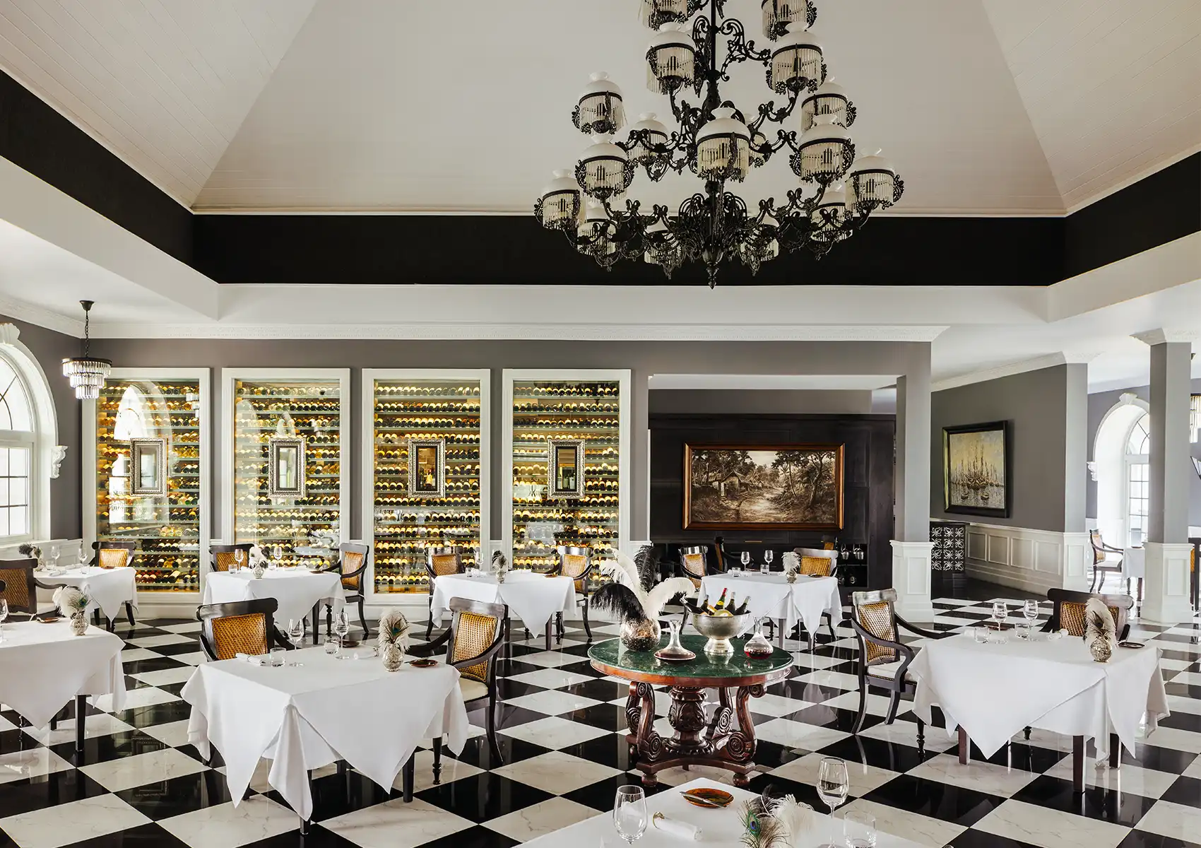 A luxurious restaurant interior featuring a black and white checkered floor, a large chandelier, white-clothed tables, and a massive illuminated wall of wine cellars, offering the perfect atmosphere for wine pairing fine dining Bali.