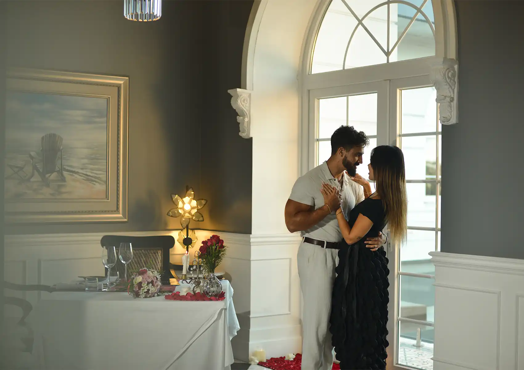 A couple embracing romantically by a large arched window, with a beautifully decorated candlelit table set for an intimate anniversary dinner Ubud in the background.