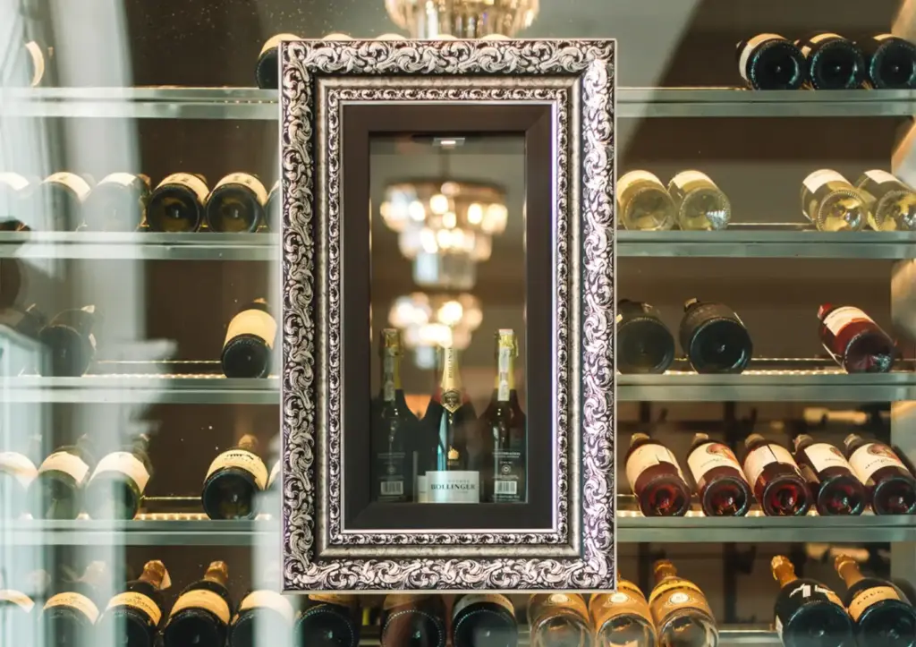 A brightly lit, glass-enclosed wine cellar showcasing an extensive collection of bottles, featuring an ornate frame highlighting premium champagne from a top wine list Bali has to offer.