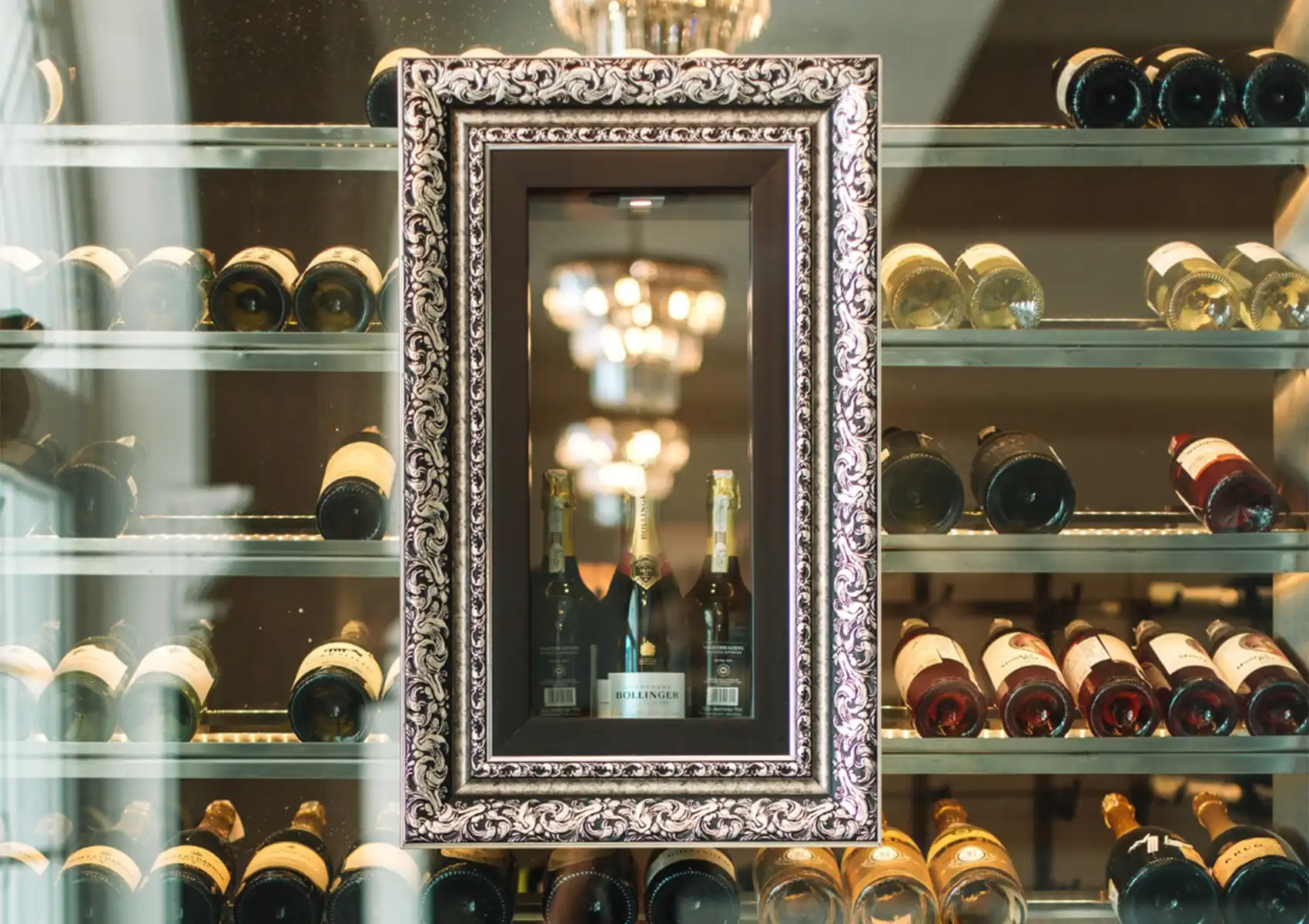 A brightly lit, glass-enclosed wine cellar showcasing an extensive collection of bottles, featuring an ornate frame highlighting premium champagne from a top wine list Bali has to offer.