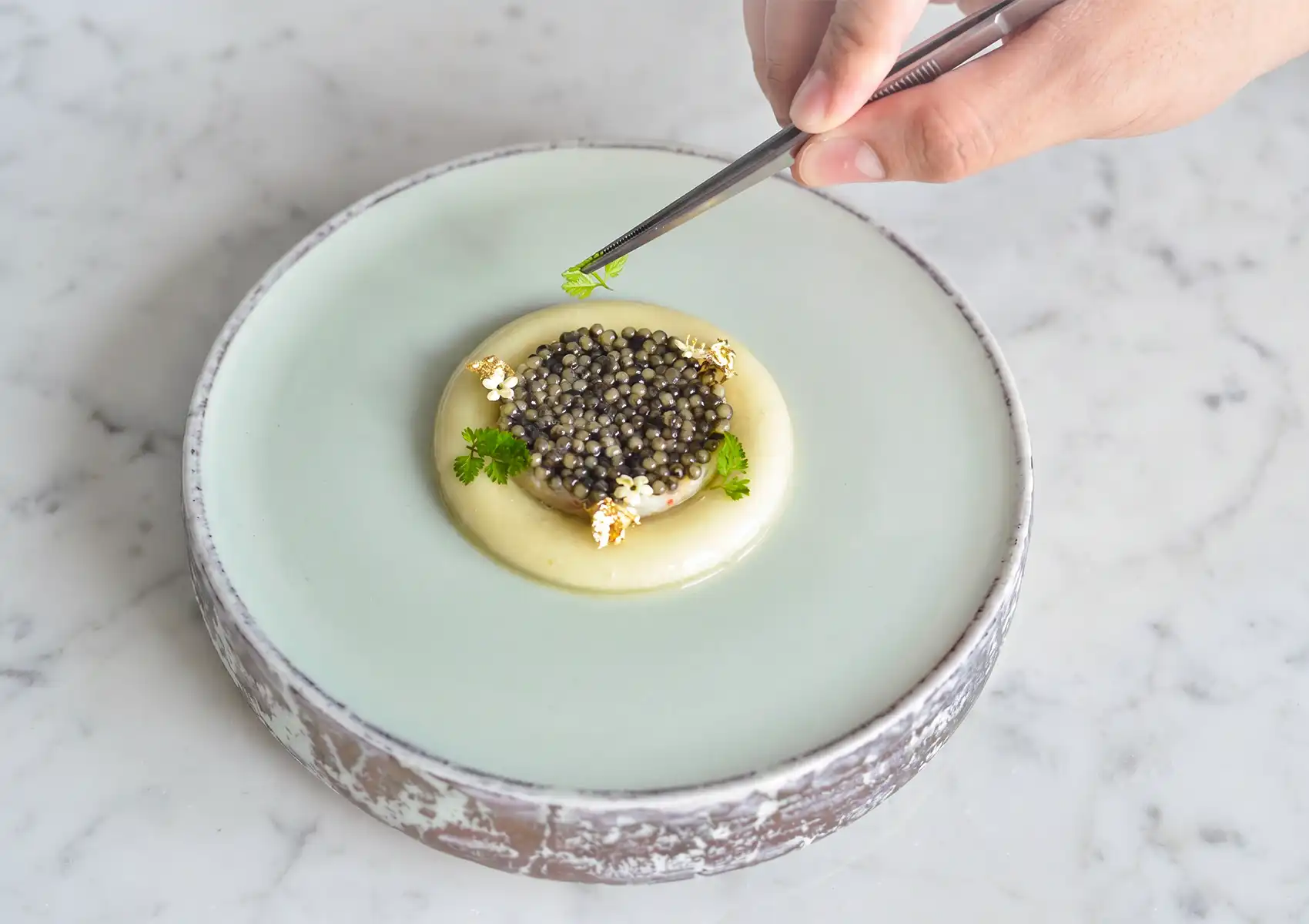 A chef using tweezers to carefully place a micro-herb garnish on a luxurious caviar dish accented with edible gold leaf, exemplifying the meticulous presentation of fine dining Ubud.