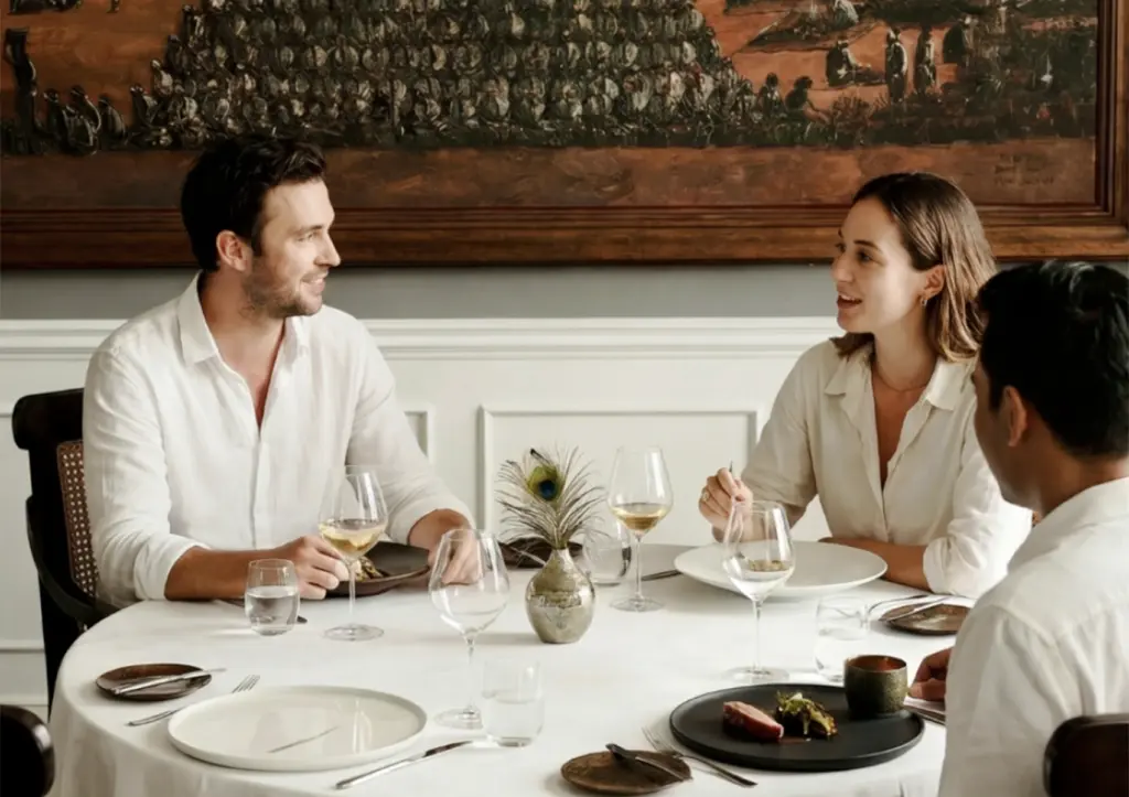 A group of friends conversing at an elegant restaurant table, demonstrating an appropriate fine dining dress code Bali with their crisp, smart-casual white shirts.
