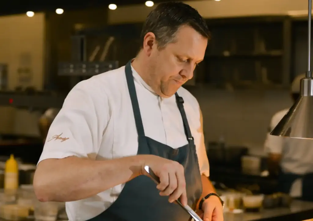 A professional chef in a white uniform and apron carefully preparing a dish in the kitchen of a fine dining Ubud restaurant.