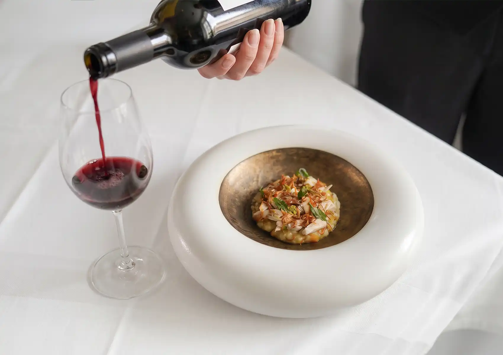 A server pouring red wine into a glass next to an elegantly plated gourmet dish on a crisp white tablecloth, showcasing a perfect pairing from a curated wine list Bali.