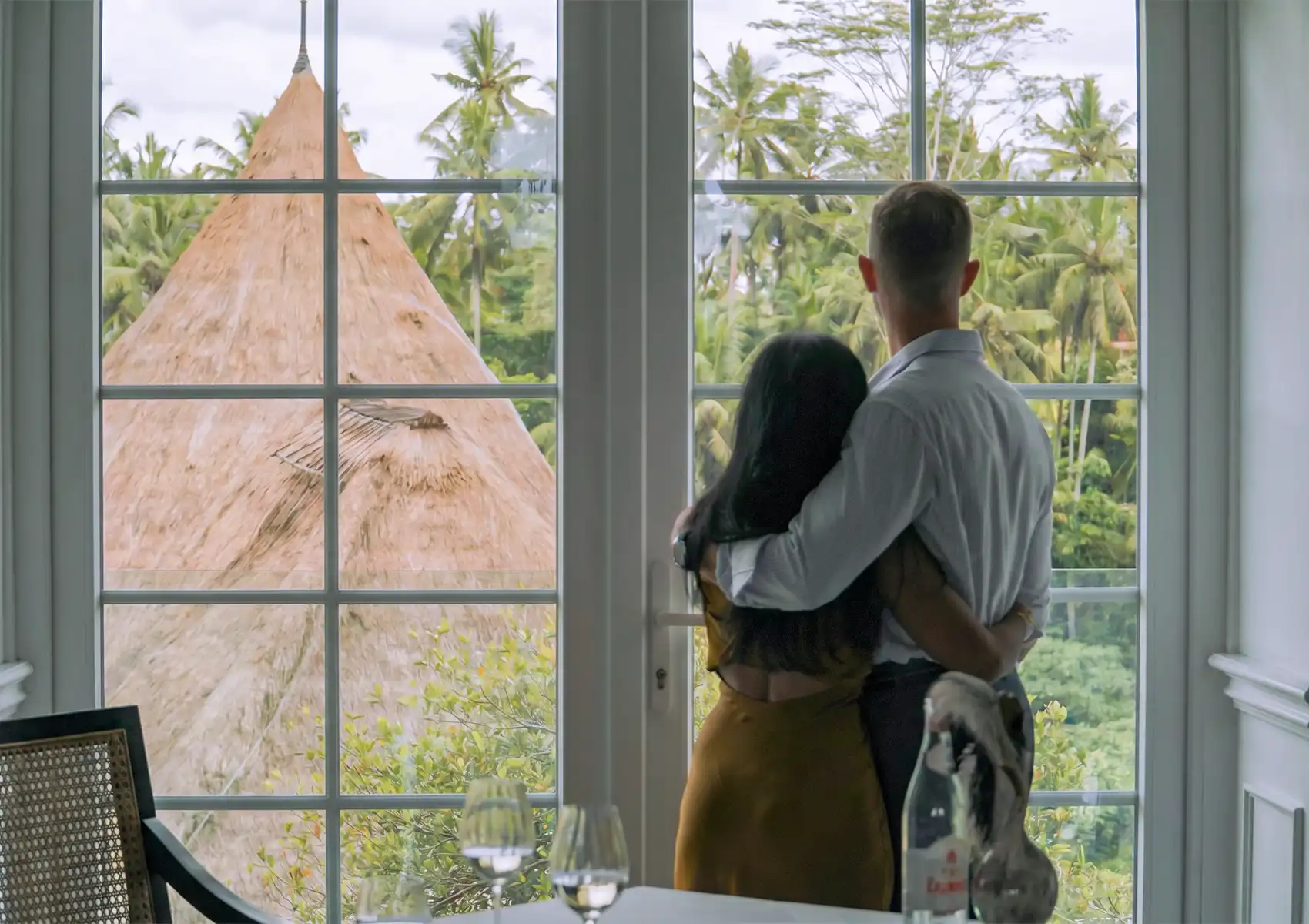 A couple embracing while looking out a large window at a lush tropical landscape and traditional thatched roof, enjoying a romantic dining experience with selections from a premium wine list Bali.