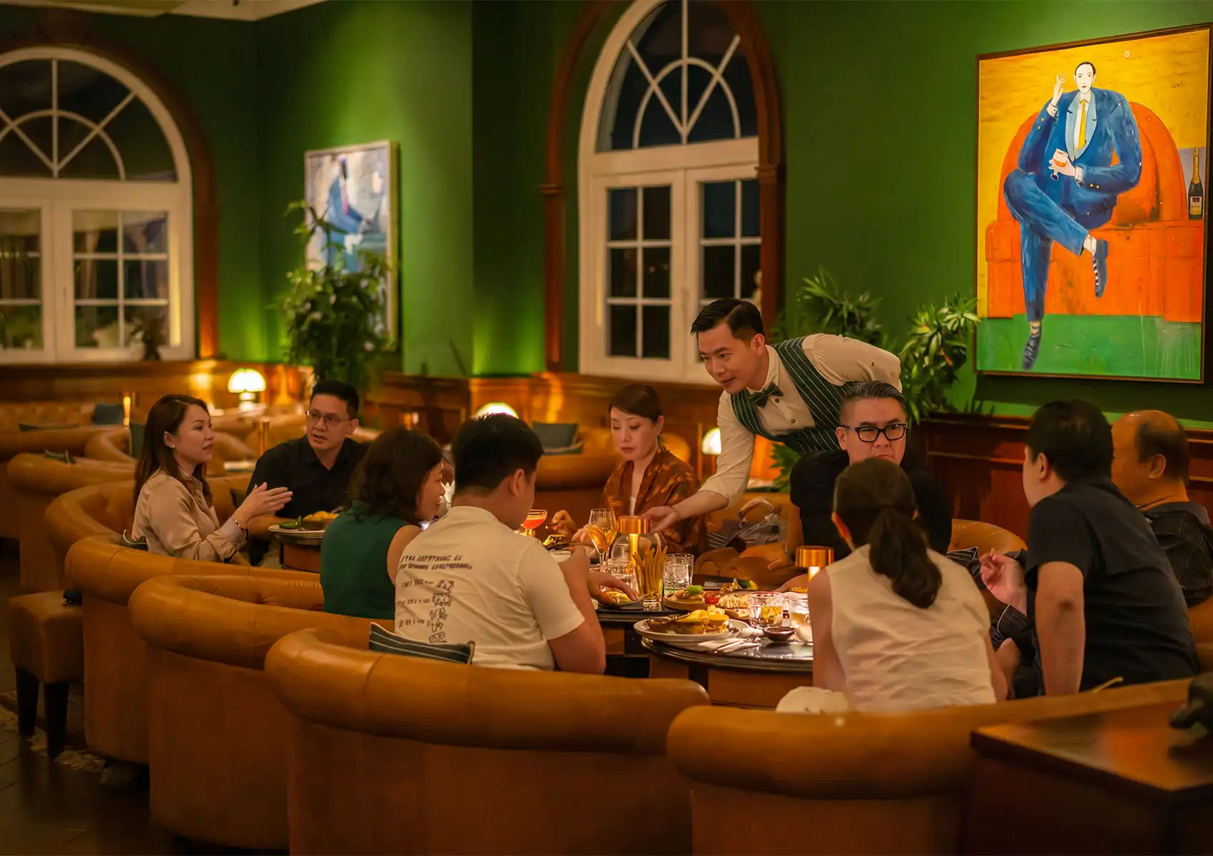 A waiter serving a group of friends relaxing in a plush leather booth inside a stylish green-walled lounge, gathering for drinks before a birthday dinner in Ubud.