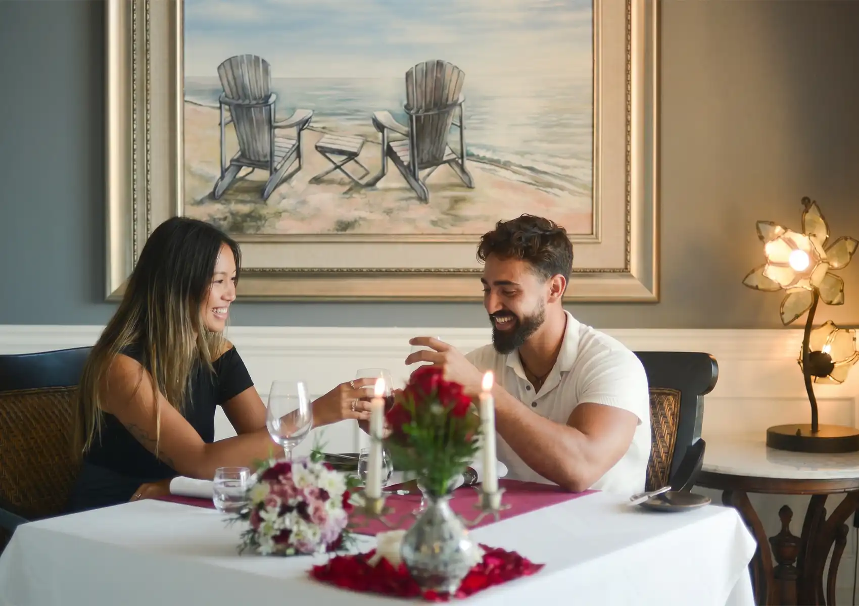 A happy couple smiling and toasting with glasses of wine across a table decorated with red roses and candles during a romantic anniversary dinner Ubud.
