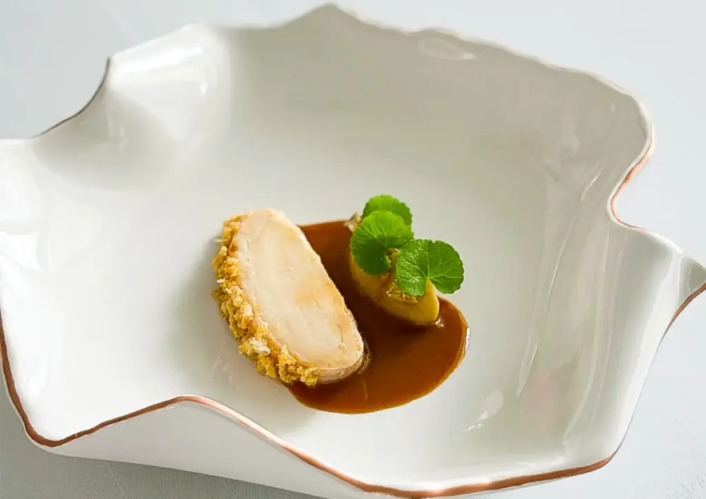 An artistic white bowl presenting a refined slice of crusted meat resting in a savory sauce with a vibrant green garnish, representing a beautifully crafted tasting menu Ubud.