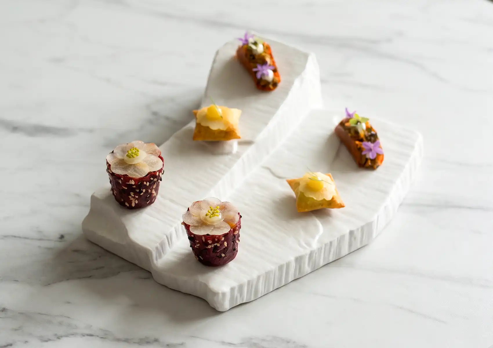 Intricately garnished canapés topped with delicate edible flowers and precise layers, arranged on a textured white stone platter on a marble table to highlight the artistry of fine dining ubud