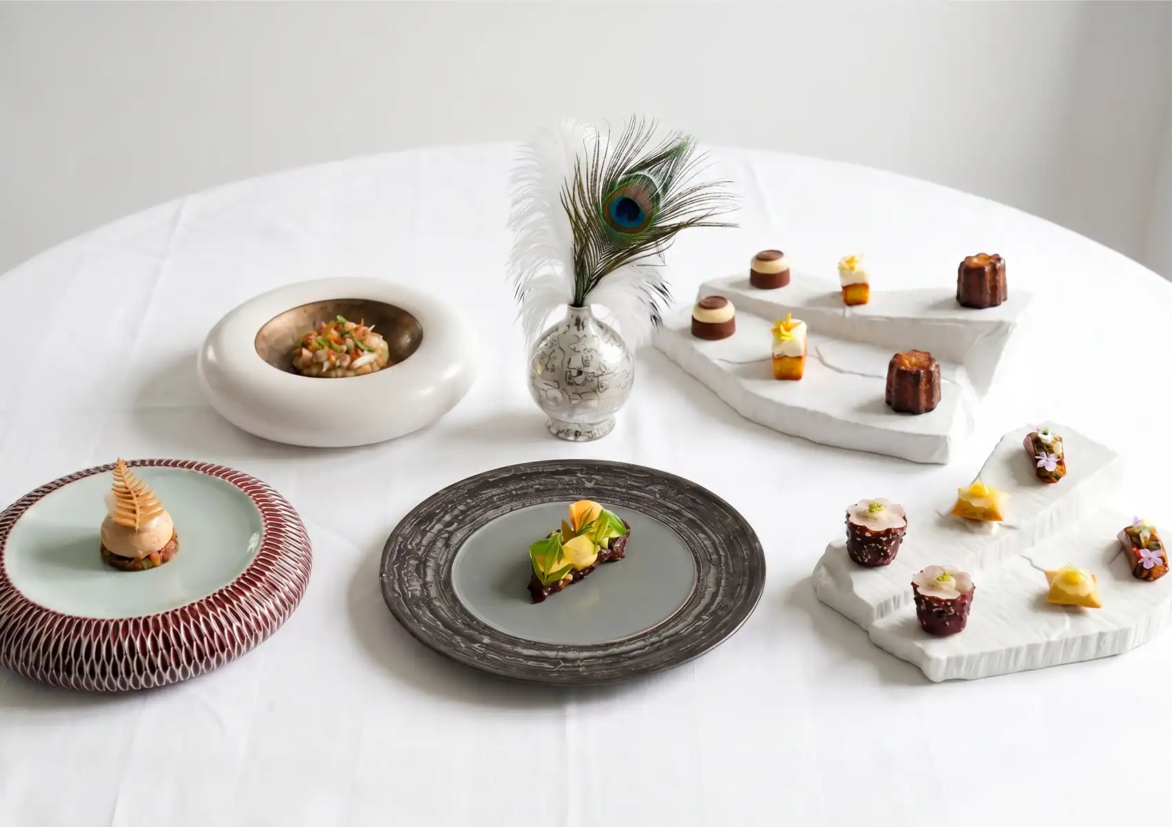 A table spread of an exquisite degustation menu featuring intricate small courses, delicate desserts on tiered white stands, and unique ceramic plates arranged around a peacock feather centerpiece, showcasing fine dining ubud.