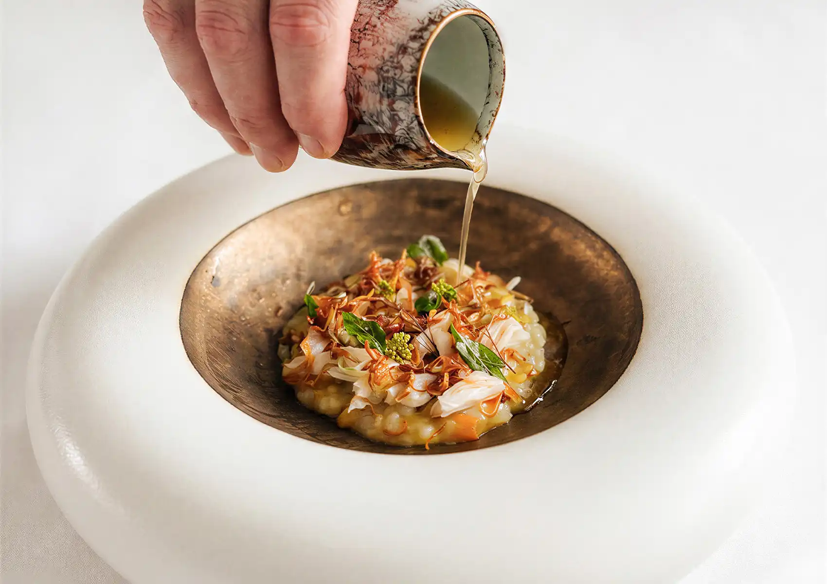 Hot broth being poured from a ceramic pitcher over a delicate crab and herb dish, showcasing the culinary artistry of fine dining Bali.