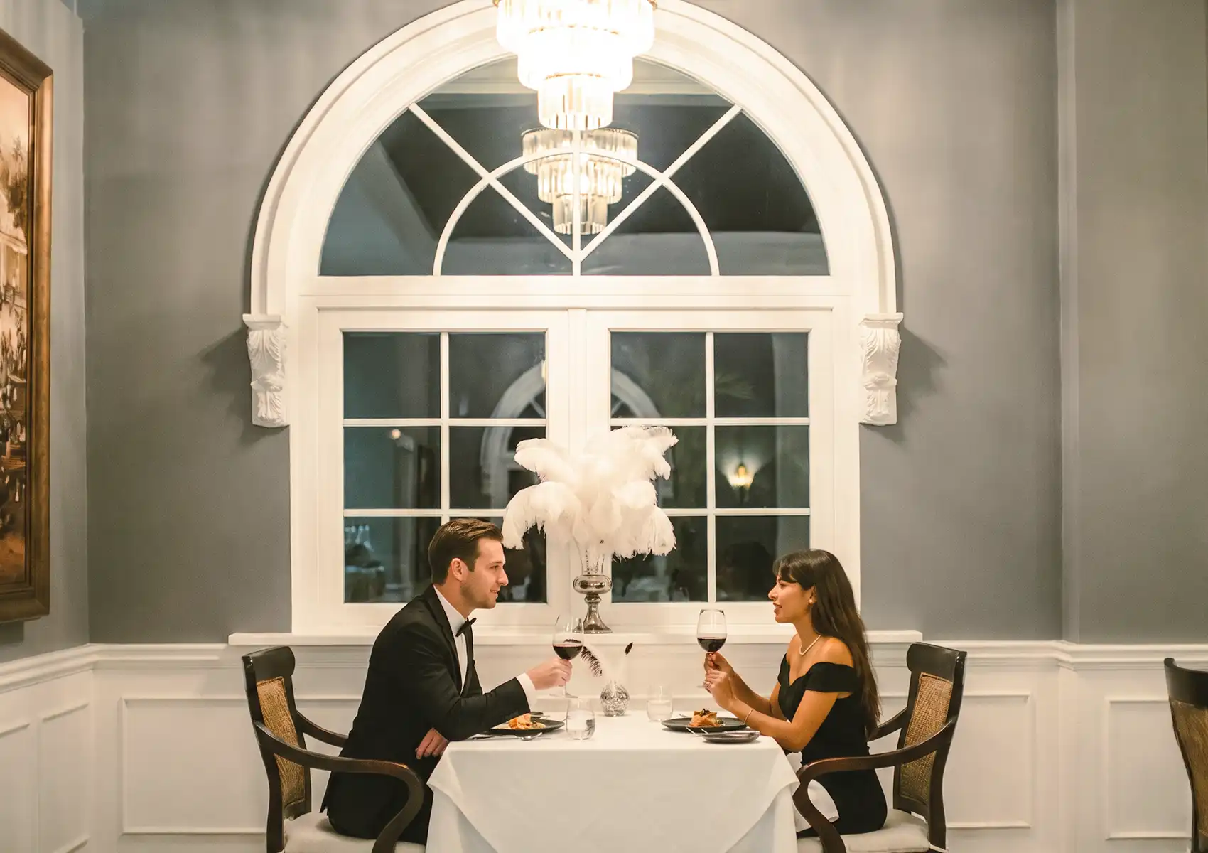 An elegant couple in formal attire toasting with glasses of red wine at a romantic white-clothed table beneath a crystal chandelier, experiencing fine dining Ubud.