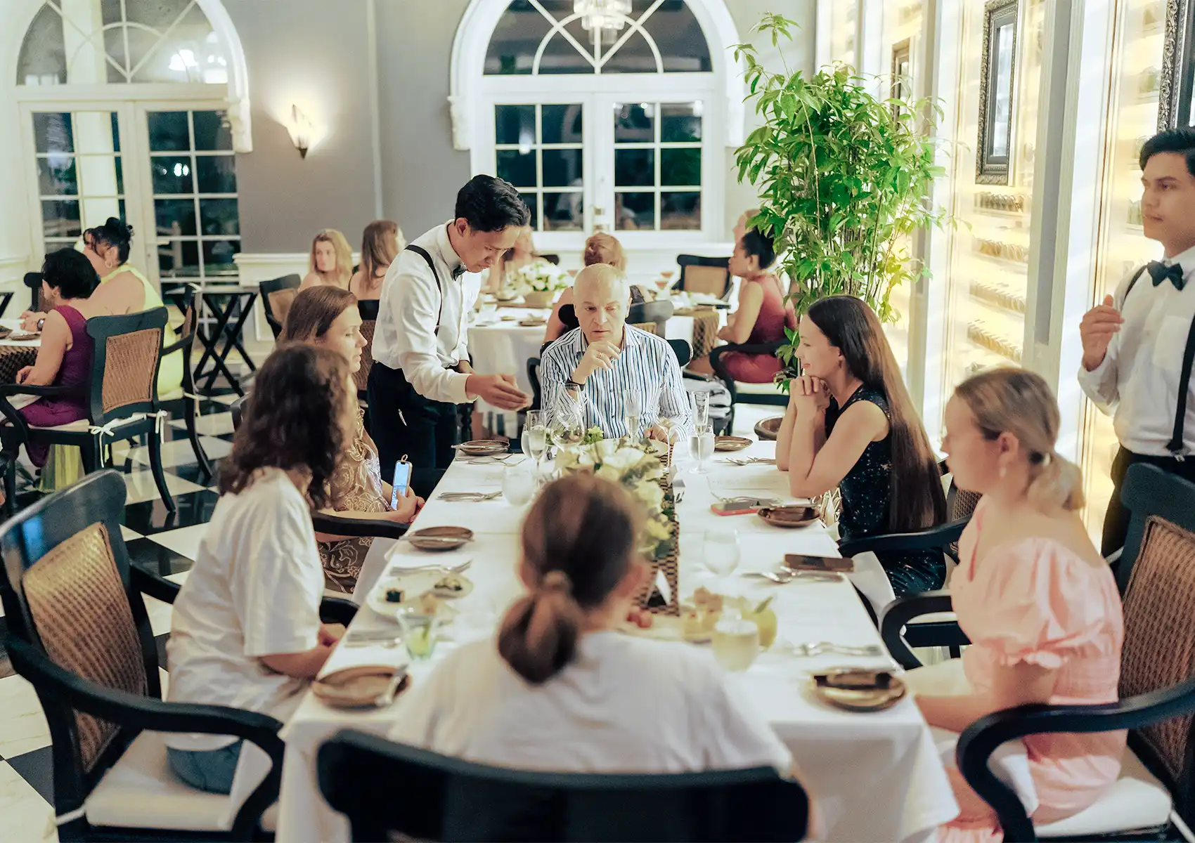A large group of guests gathered around a long white-clothed table being served by professional waitstaff in a bright, elegant restaurant setting, capturing the atmosphere of fine dining ubud.
