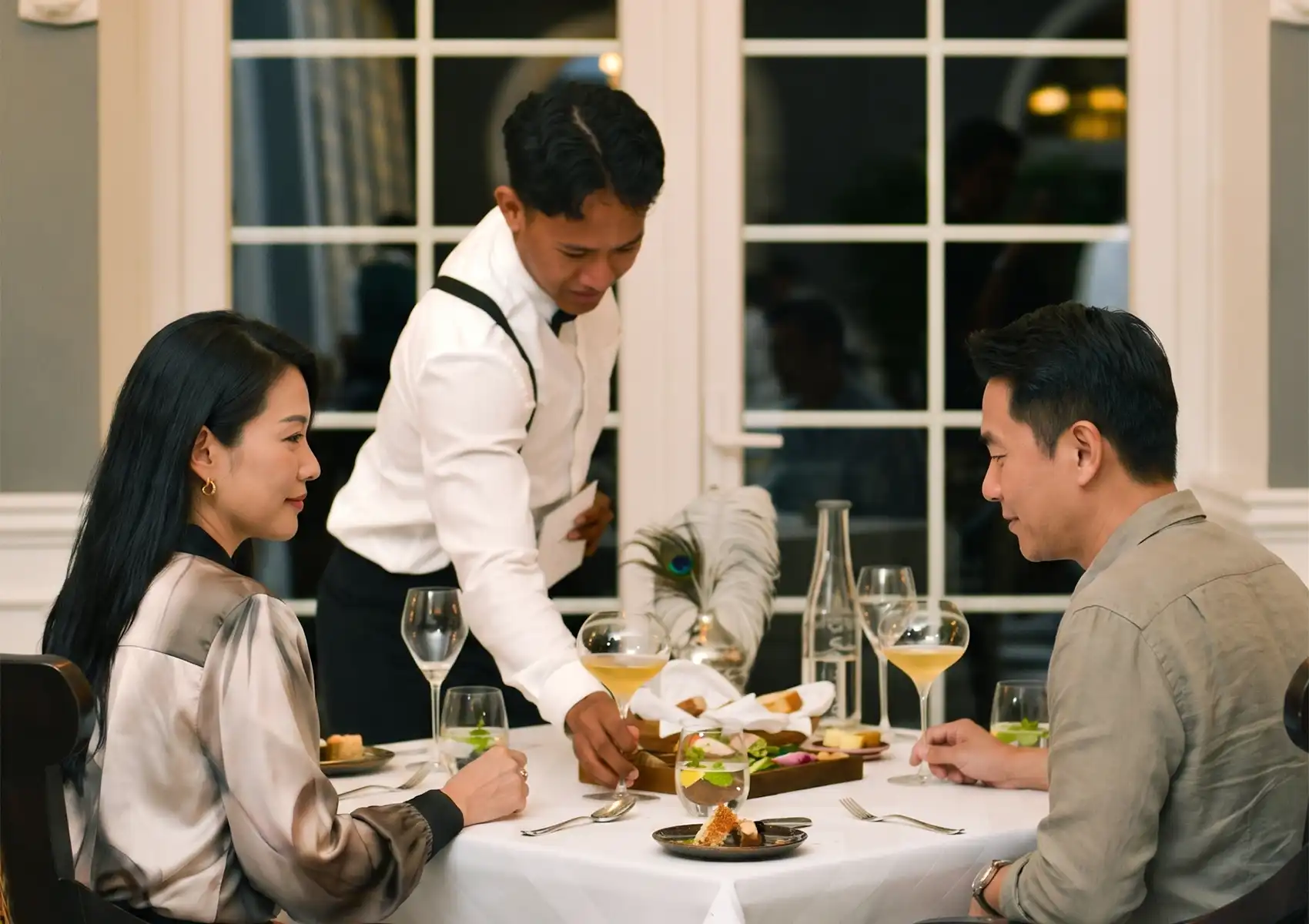 A professional waiter in suspenders serves a smiling couple enjoying wine and a beautifully presented meal at a white-clothed table, capturing a romantic evening of fine dining ubud.