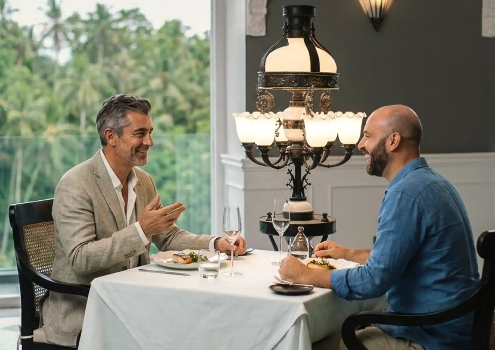 Two men laughing and conversing during a meal at an elegant white-clothed table next to an ornate vintage lamp, enjoying an experience of fine dining ubud with a lush jungle view.