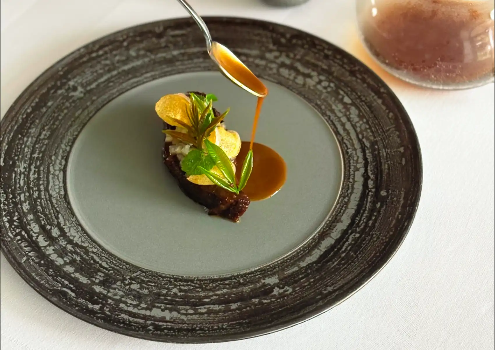 A close-up of a rich sauce being poured from a spoon over an elegantly plated dish garnished with fresh leaves and crisps on a textured ceramic plate, representing the culinary artistry of fine dining ubud.